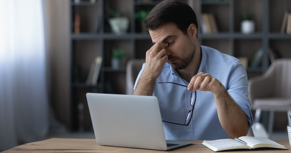 Adult sitting at a desk with a laptop and notebook in front of him. He is visibly stressed or exasperated, holding his glasses in one hand and rubbing the bridge of his nose with the other.