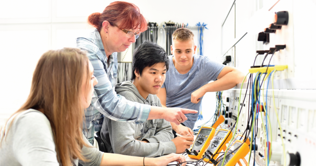 A woman with red hair points at a multimeter as three students watch attentively in a technical training environment. The workbench is filled with cables and electronic tools.